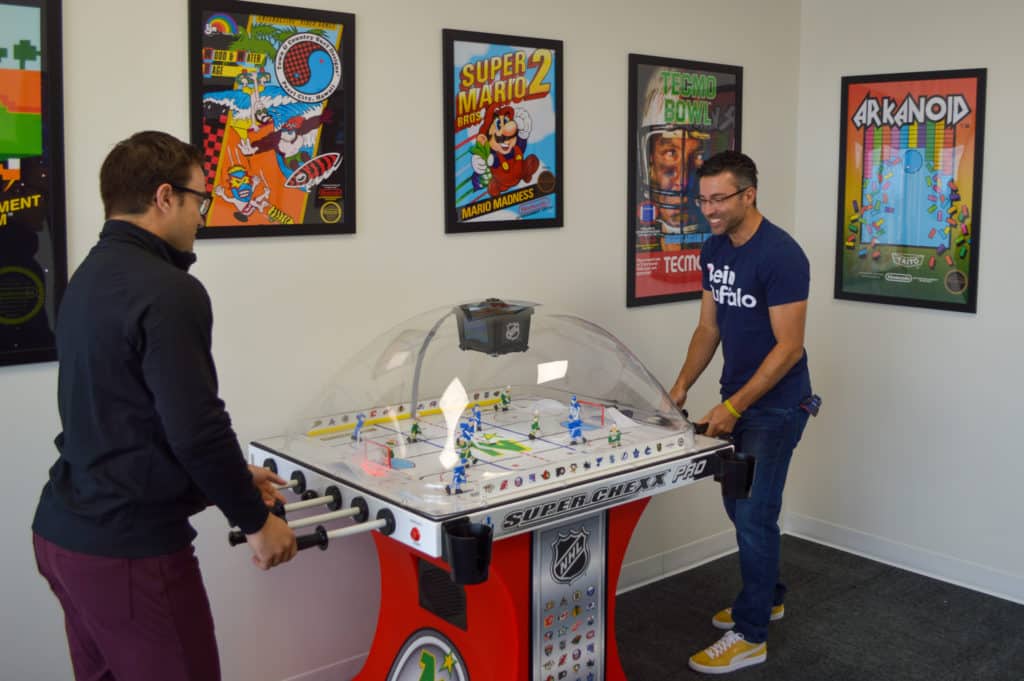 Two men stand at either side of a bubble hockey table playing hockey. Background is white walls with fun 80s themed game posters