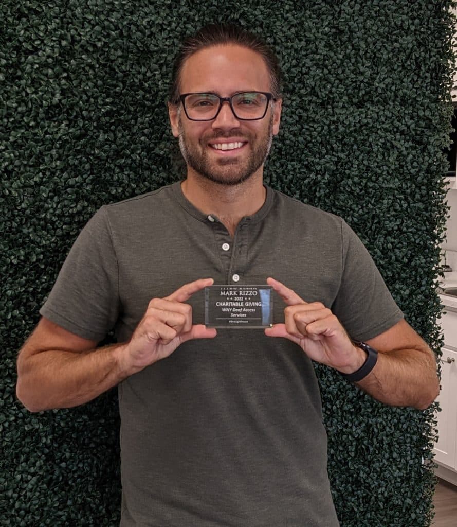 Man wearing a green shirt in front of a green background, holding up a plaque in honor of his support of WNY Deaf Access Services